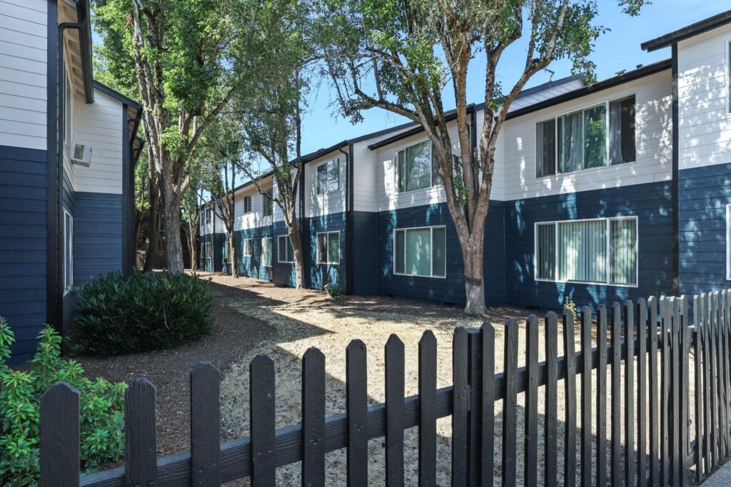 Apartment complex exterior with blue and white buildings, surrounded by tall trees and a wooden fence.