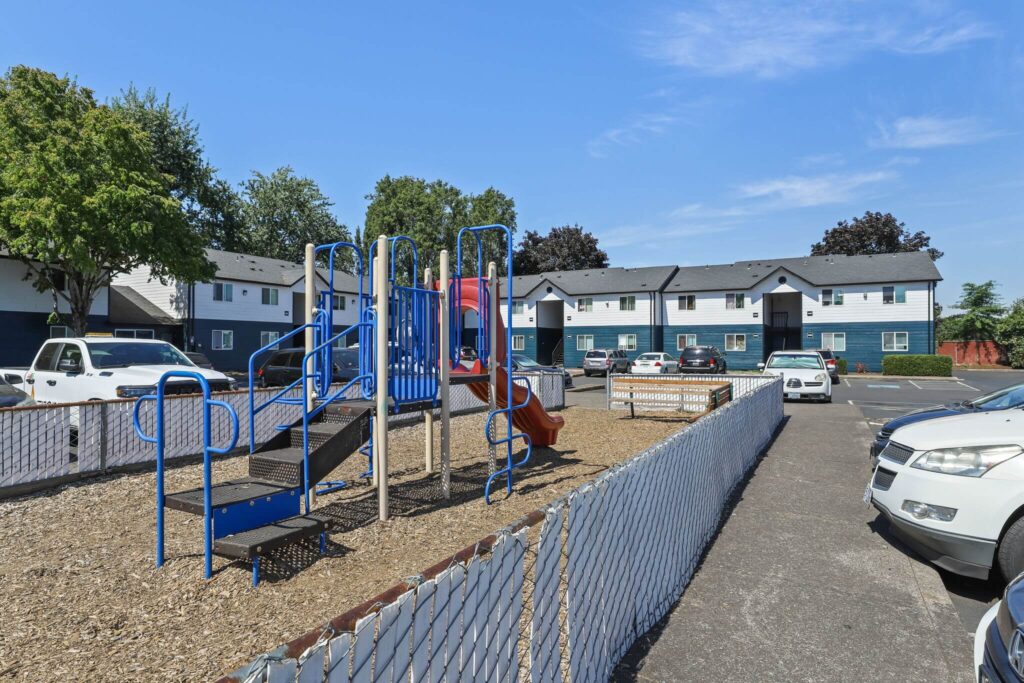Fenced playground with woodchips, red slide, and benches, surrounded by parked cars and residential apartments.