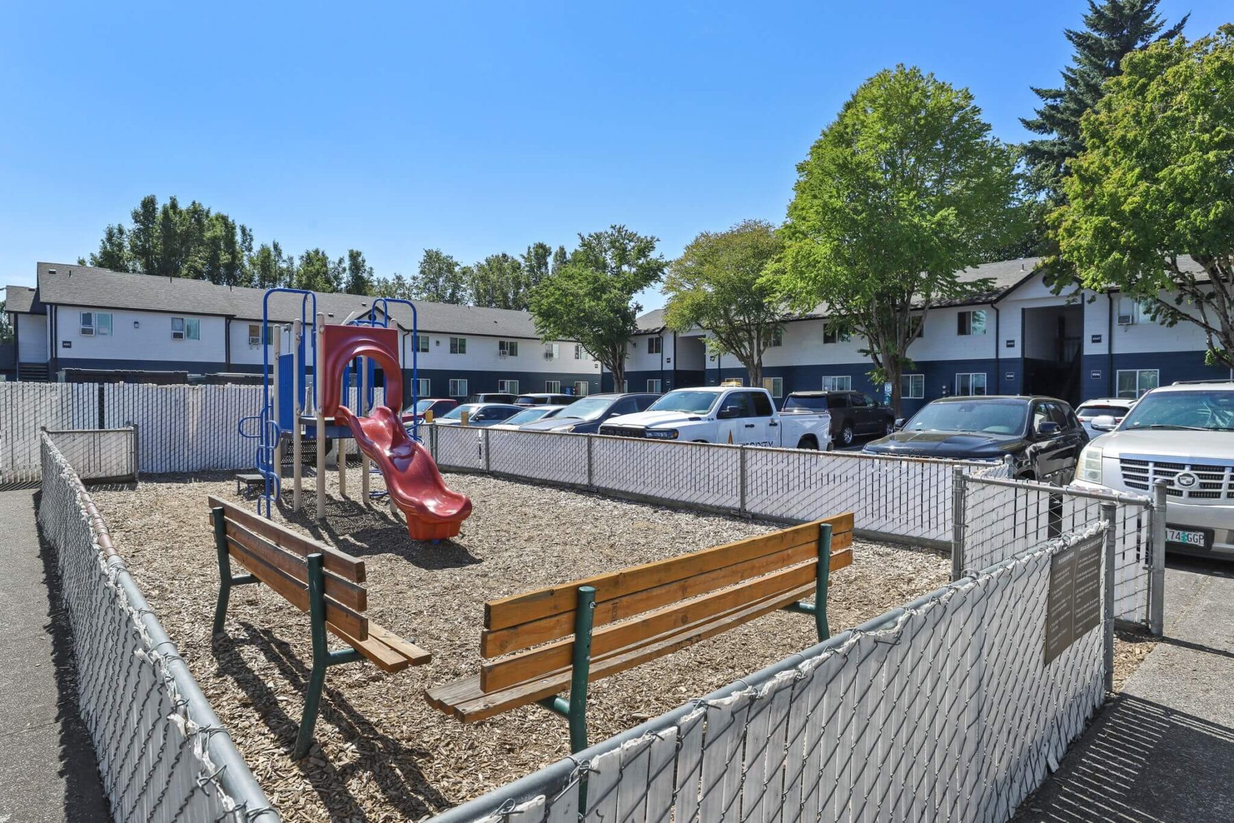 Fenced playground with woodchips, red slide, and benches, surrounded by parked cars and residential apartments.