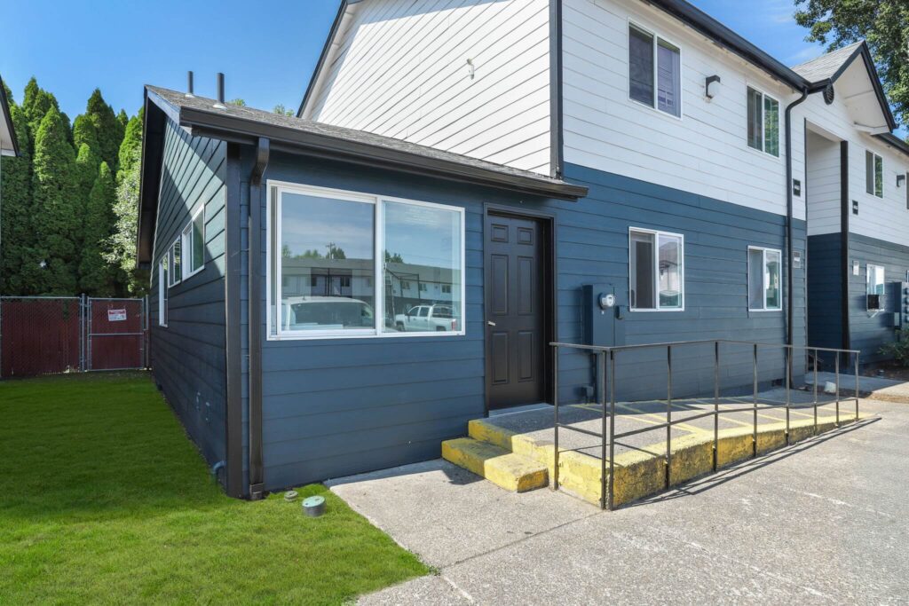 A two-story house with dark blue siding on the lower half and white on the upper, featuring a ramp and steps leading to a dark door, with a well-kept lawn.