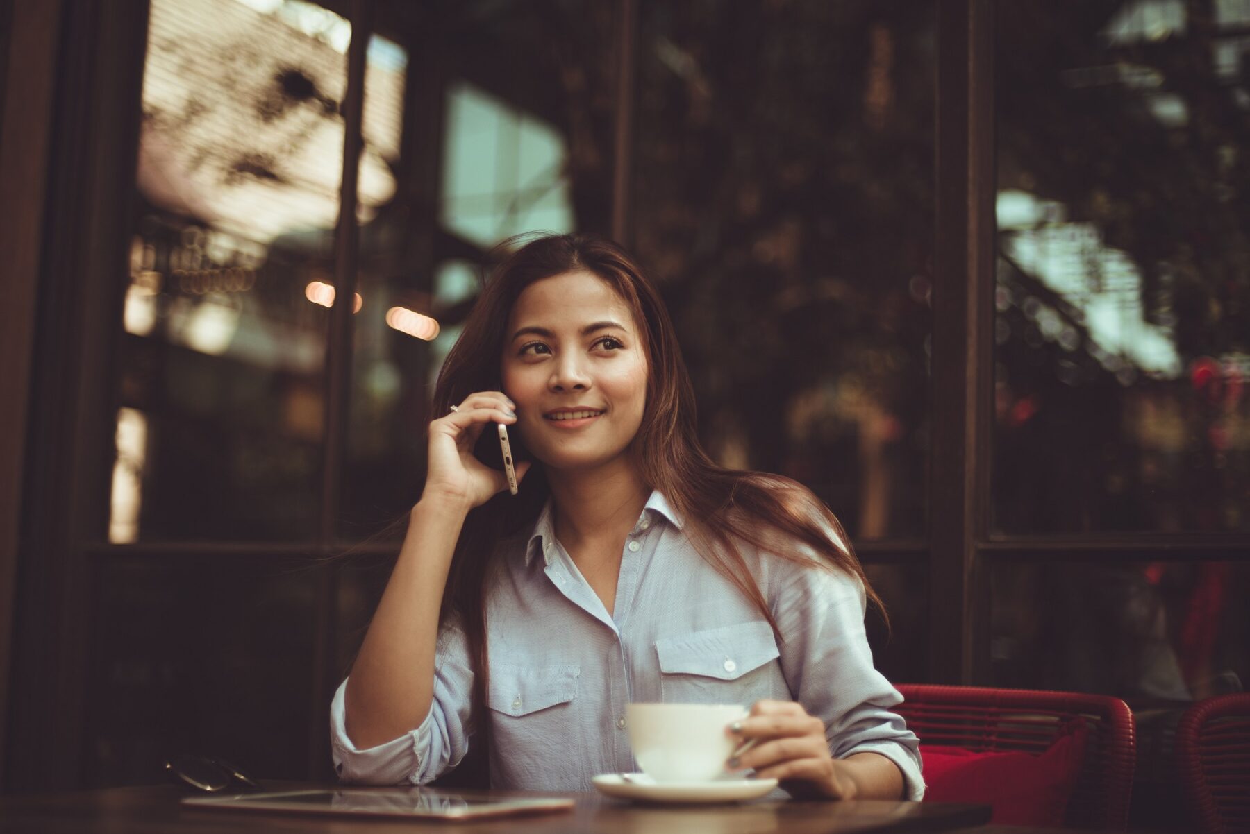 A woman with long hair sits at a cafe table, smiling while talking on her phone and holding a cup of coffee. A tablet is on the table in front of her, and large windows are in the background.
